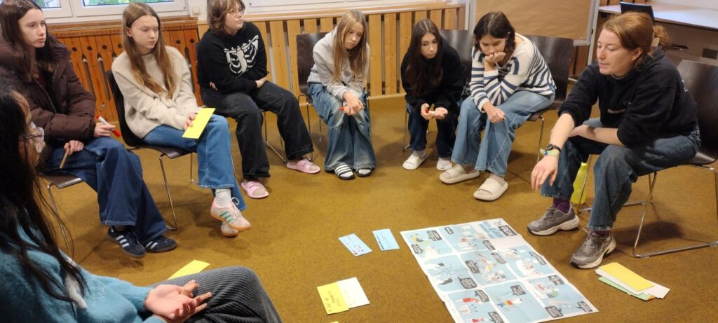 Jugendliche sitzen im Kreis auf Stühlen, schauen sich ein Poster auf dem Boden an.